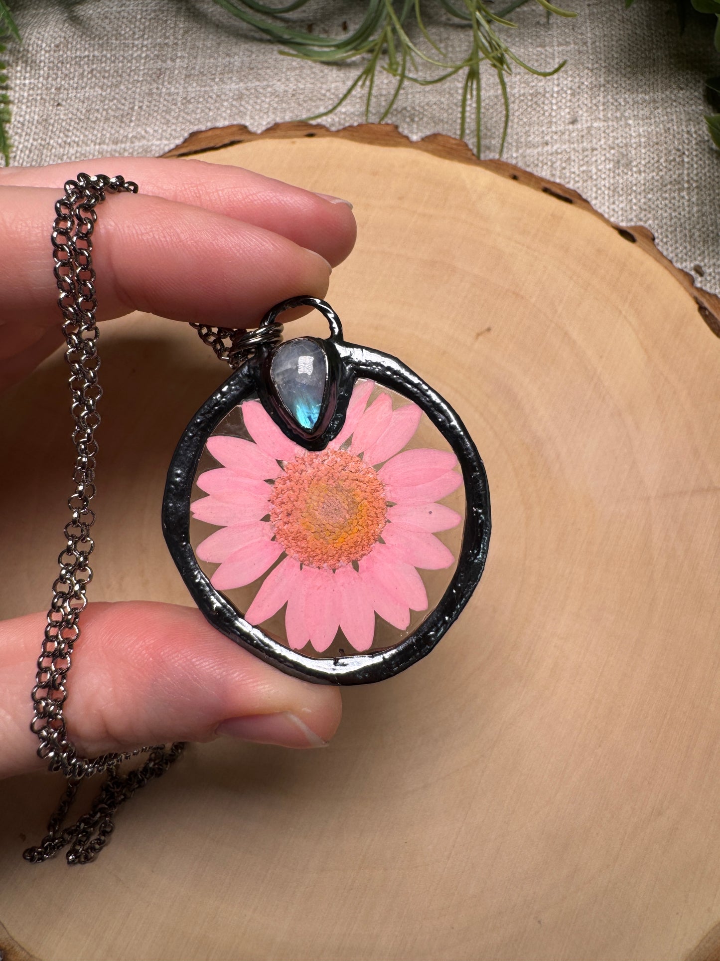 Necklace with a heart-shaped pendant featuring a pink flower, held by a hand on a wooden surface.
